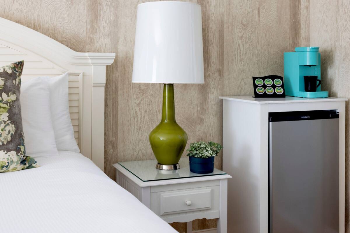 A hotel room with a bed, a nightstand with a lamp, a coffee maker, a small plant, and a refrigerator.