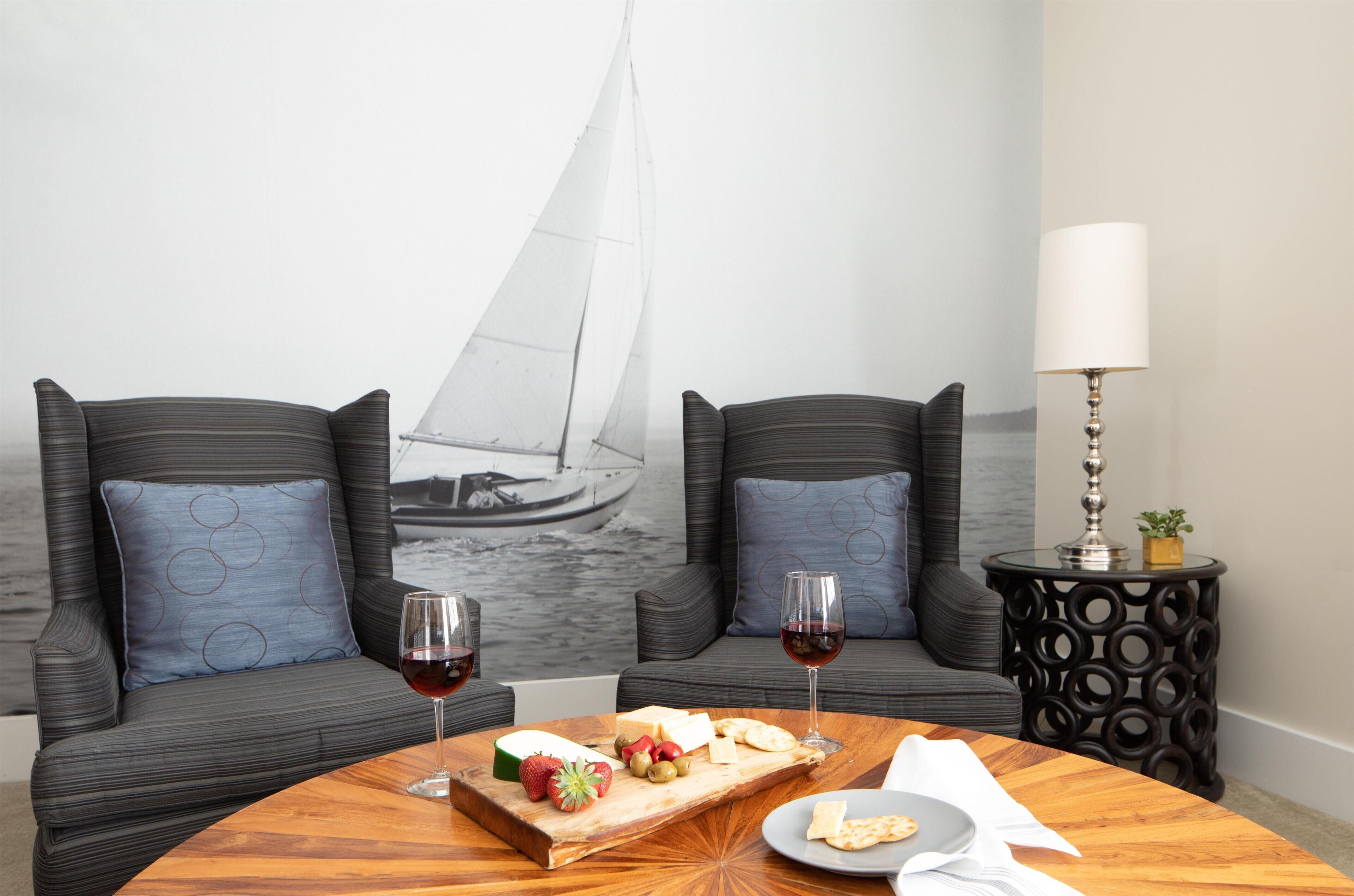 Ein Essbereich mit einem runden Holztisch, gedeckt mit einer Käseplatte, Obst und Weingläsern. Zwei gepolsterte Stühle mit gemusterten Kissen zeigen auf eine Wand mit einem Segelboot-Wandgemälde. Ein kleiner Beistelltisch mit Lampe und Pflanze ist sichtbar.