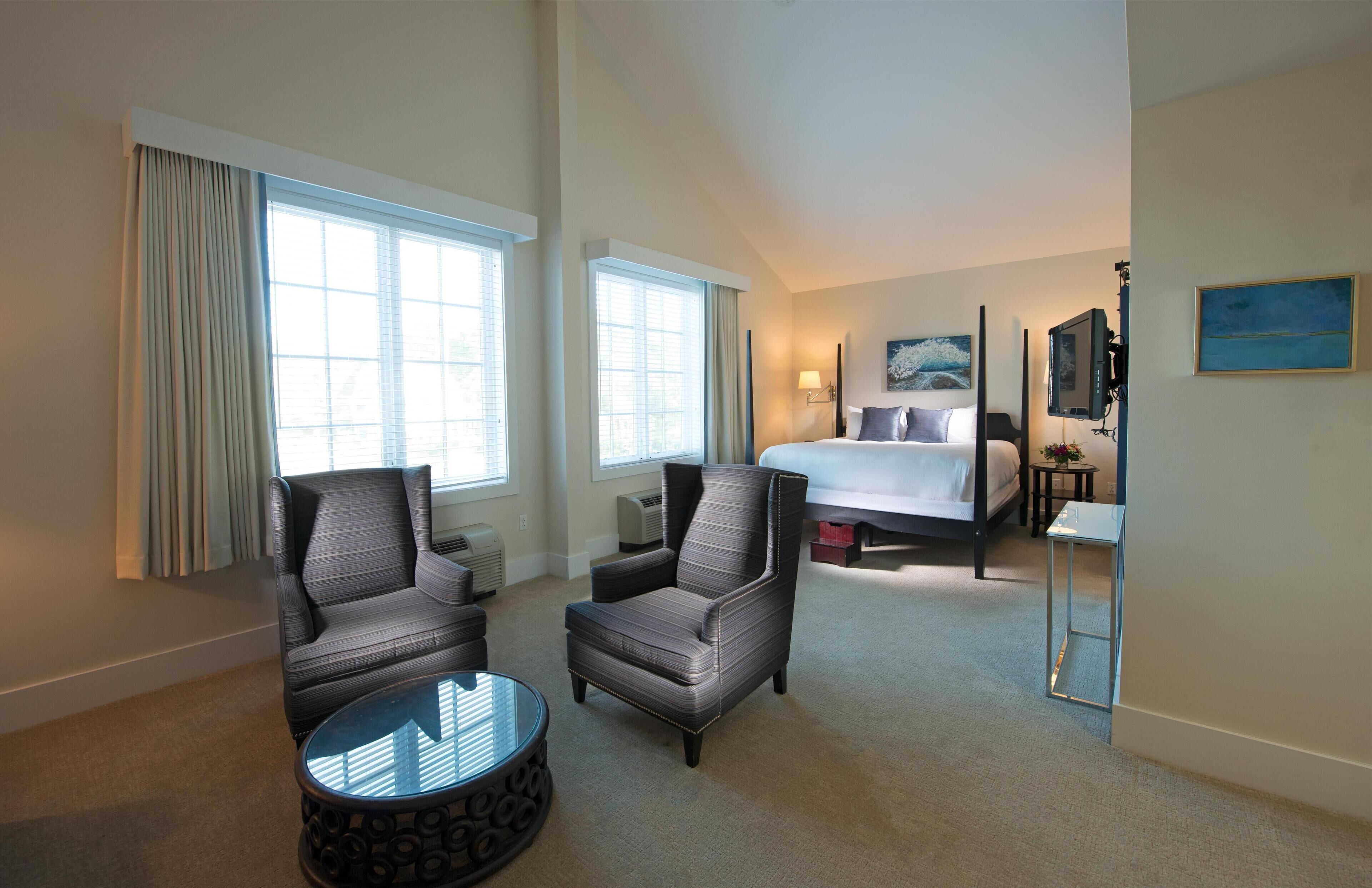 Quarto de hotel com uma cama grande, duas poltronas, uma mesa pequena e uma televisão.