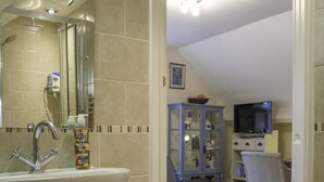 Un baño con lavamanos, espejo y ducha. En la habitación contigua hay un televisor y un armario.