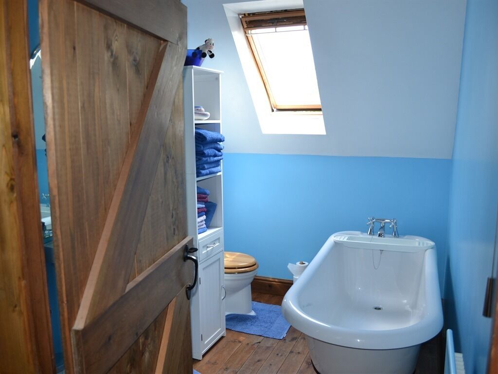 A bathroom with a wooden door, a white toilet, a blue wall, a window, and a white bathtub.