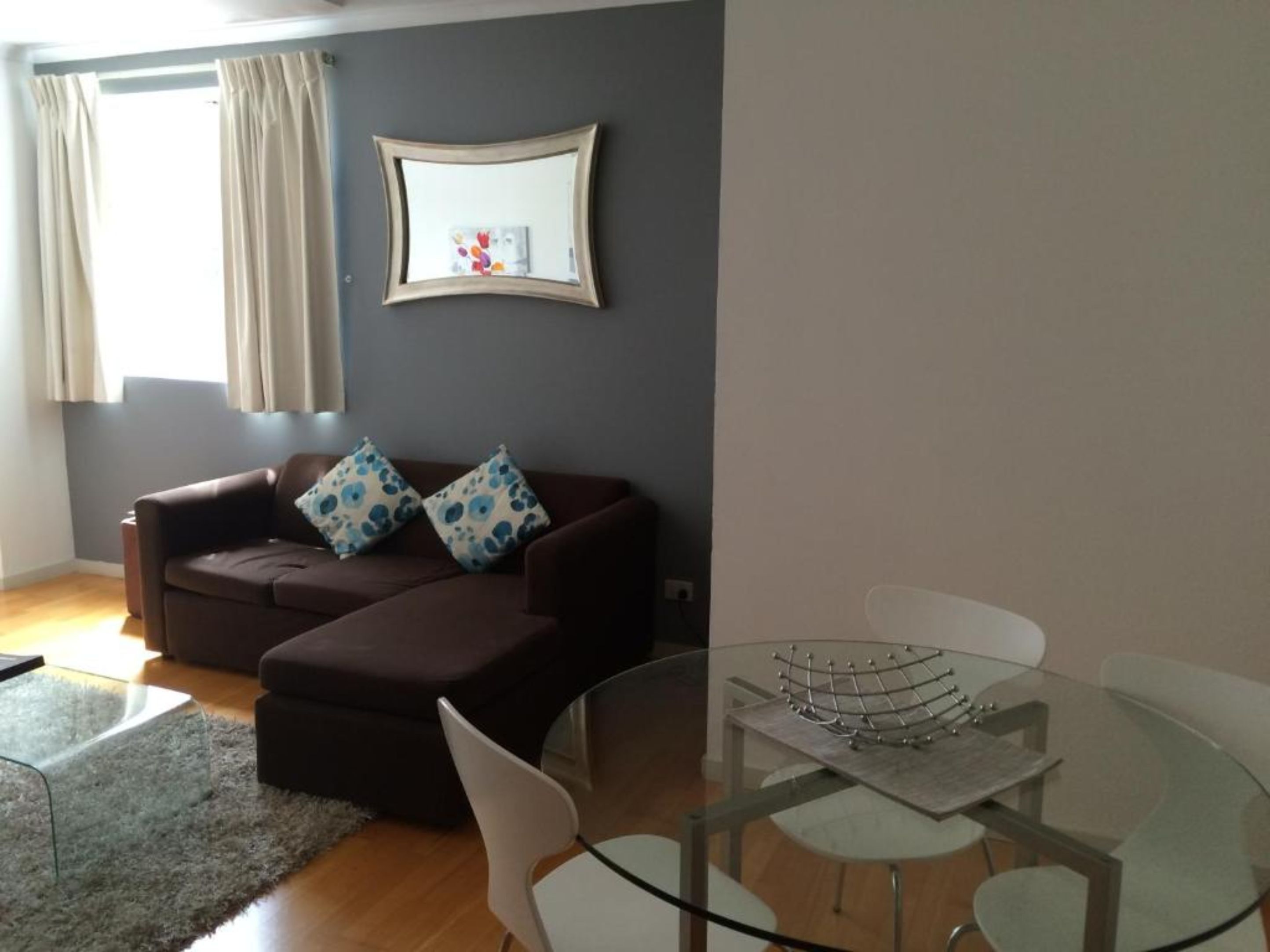 Una sala de estar con un sofá marrón, dos cojines estampados, una mesa de centro de vidrio y una silla blanca.