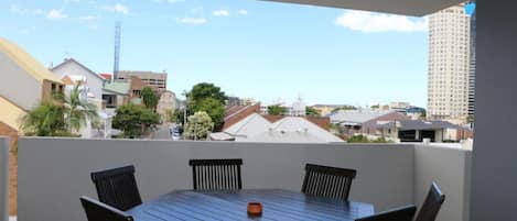 A balcony with a wooden table and chairs, overlooking a residential area with various buildings and a clear sky.