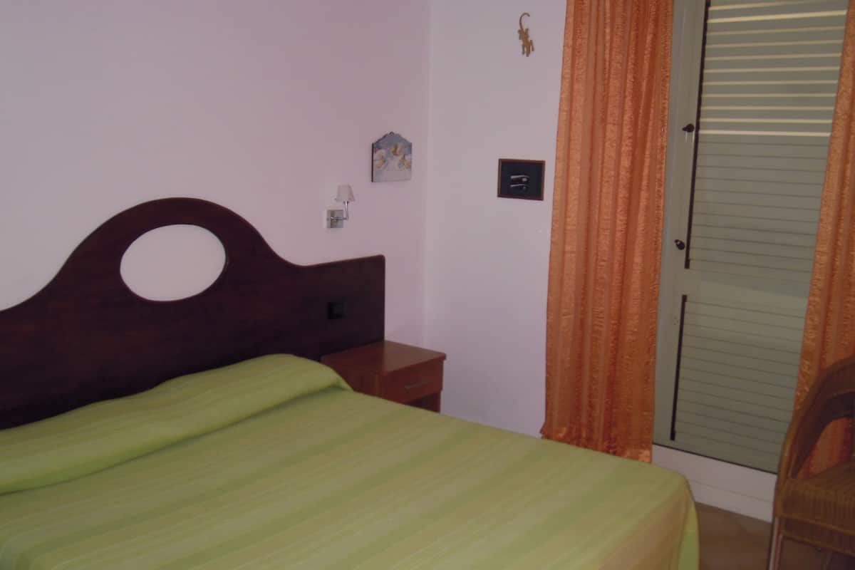 Una cama individual con cabecera de madera, una mesita de noche, un interruptor de luz en la pared y una puerta con cortina.