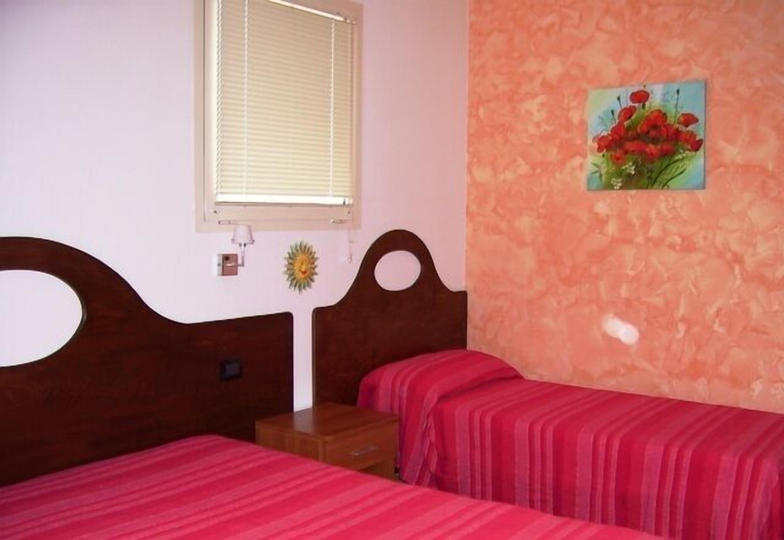 A bedroom with two beds, a window with blinds, a nightstand, a small table, and a wall-mounted picture of flowers.