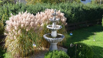 Un jardín con una fuente escalonada, diferentes arbustos y una valla de madera al fondo.