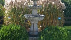 Una fuente de piedra con varios niveles, rodeada de exuberante follaje y altas hierbas ornamentales.