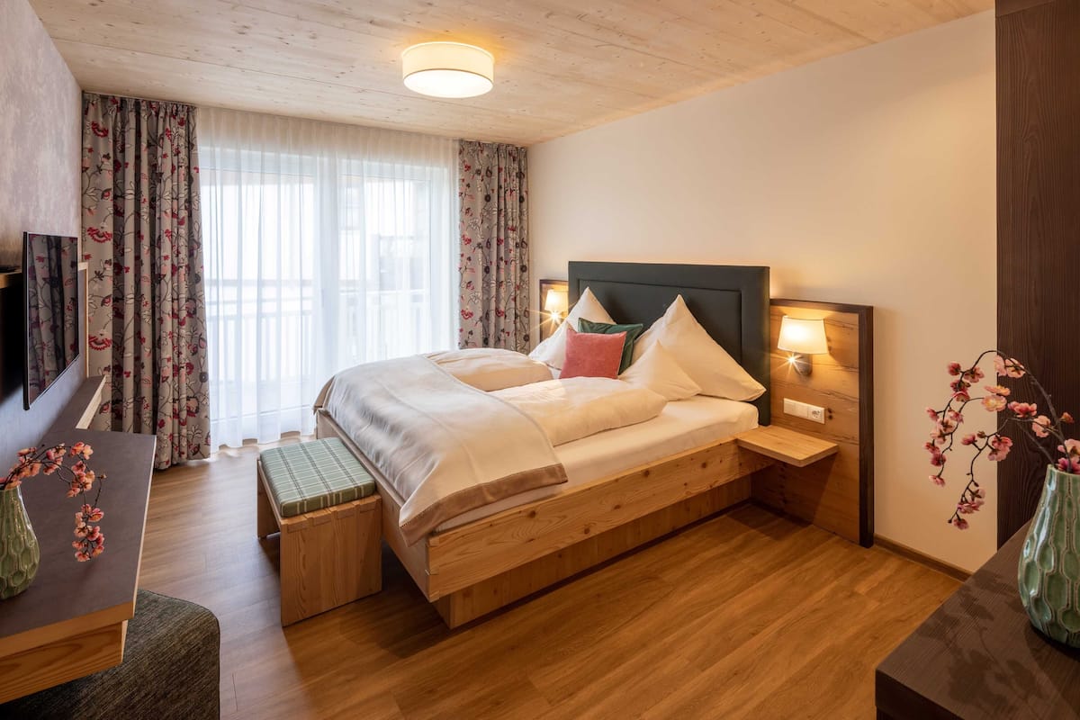 Une chambre moderne avec un lit en bois, des tables de chevet avec des lampes, un banc au pied du lit et un téléviseur sur le mur de gauche.