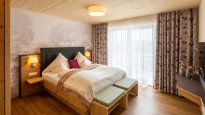 Una cama de madera con un banco, una mesita de noche con lámpara y una ventana con cortinas florales.