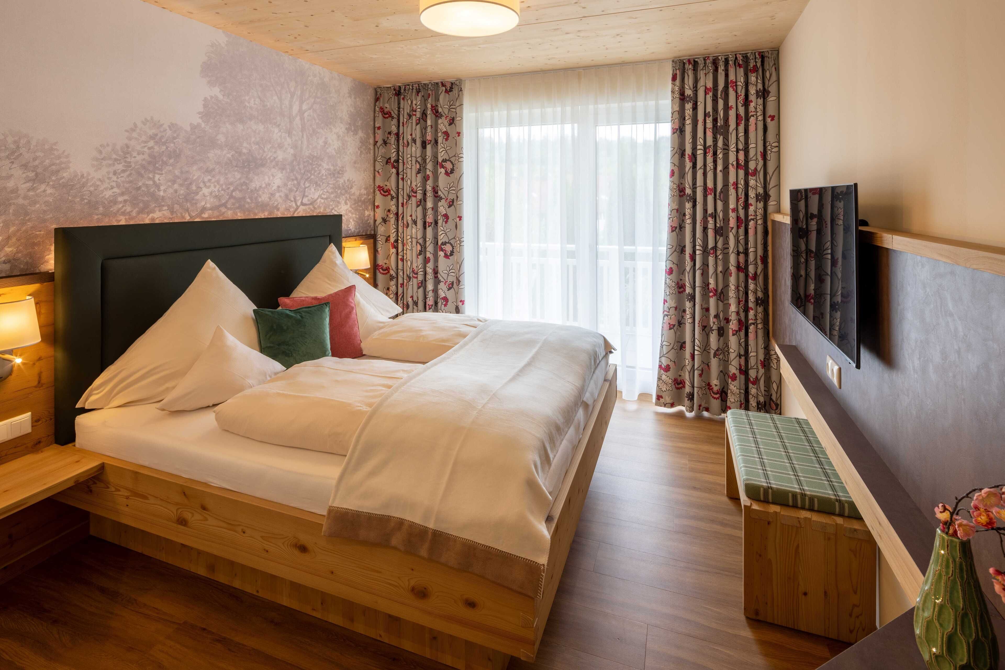 Un dormitorio con cama de madera, un banco, un televisor montado en la pared y una ventana con cortinas.