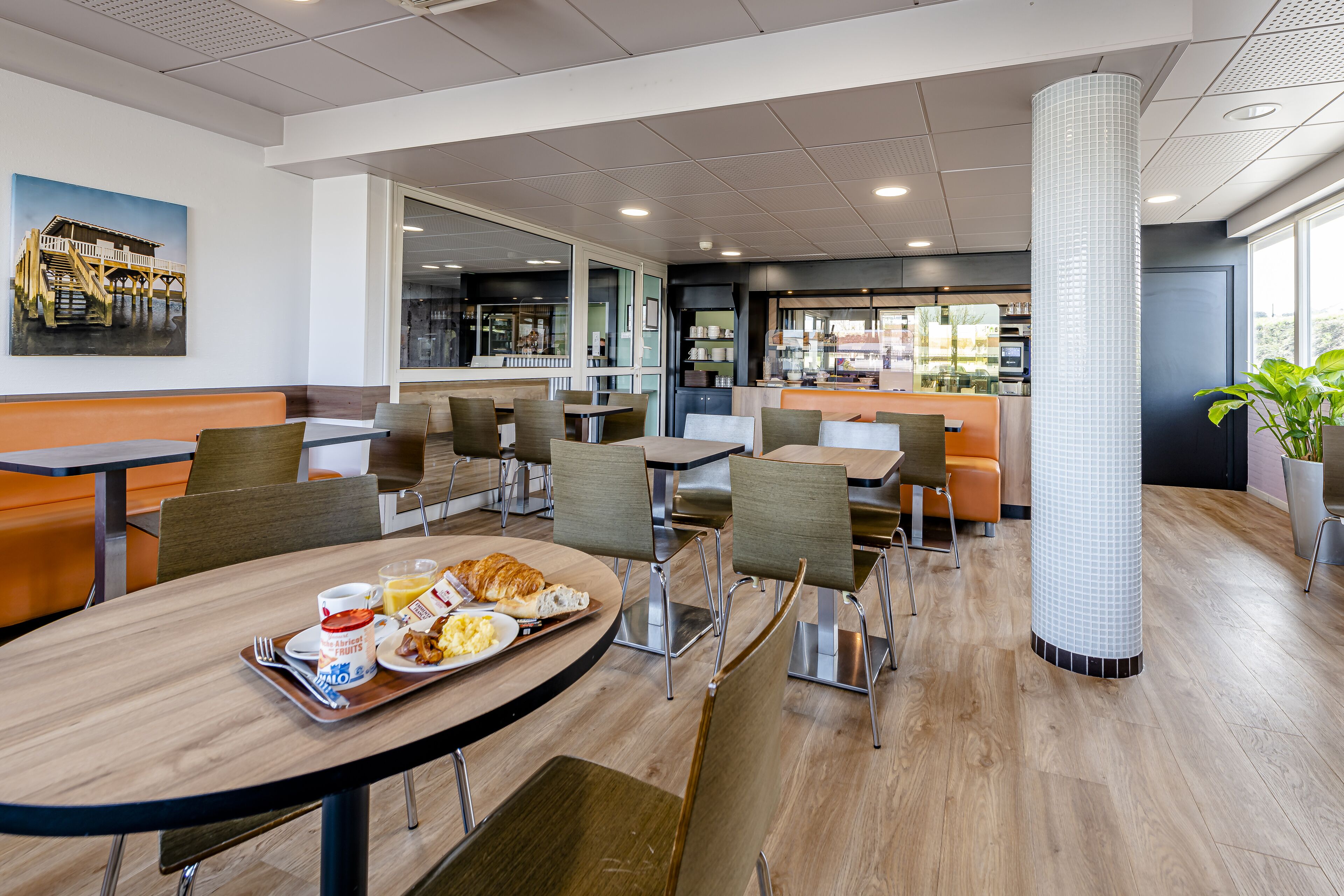 Un espace de restauration moderne, doté de plusieurs tables et chaises, d’un pilier central et offrant une vue sur la cuisine.