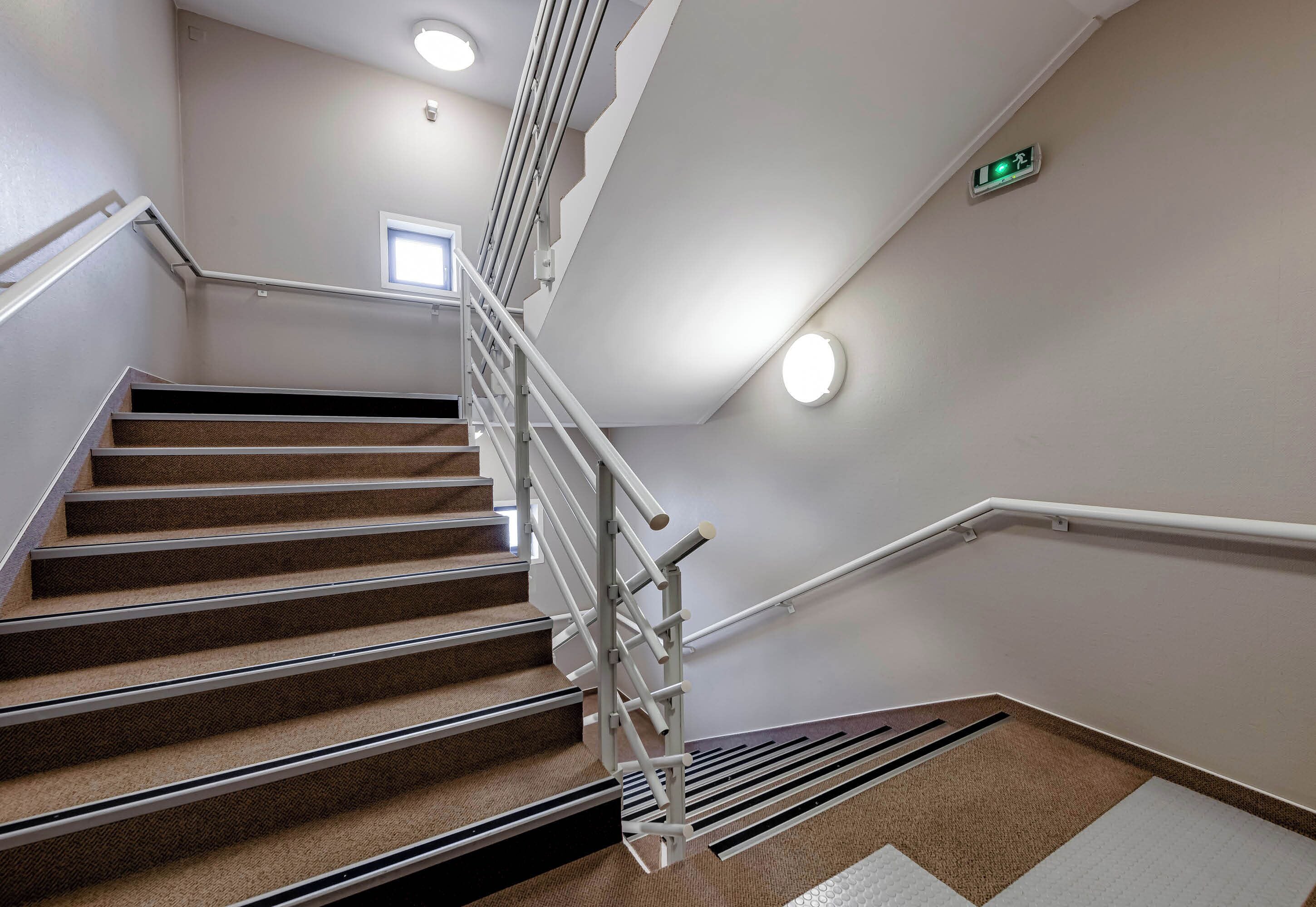 Un escalier moderne avec une rampe en métal, des marches recouvertes de moquette et une petite fenêtre en haut.