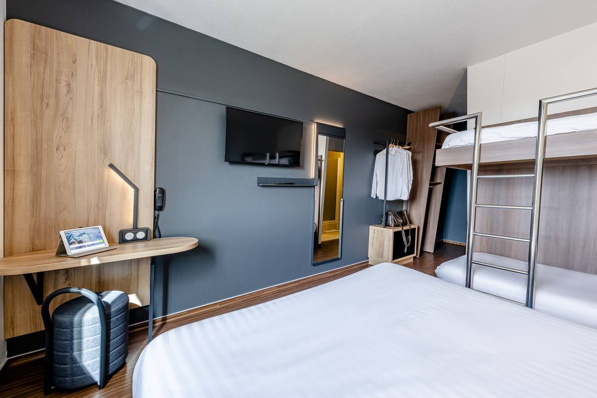 Une chambre d’hôtel moderne avec un grand lit, un bureau avec un ordinateur portable, un téléviseur fixé au mur et un lit superposé.