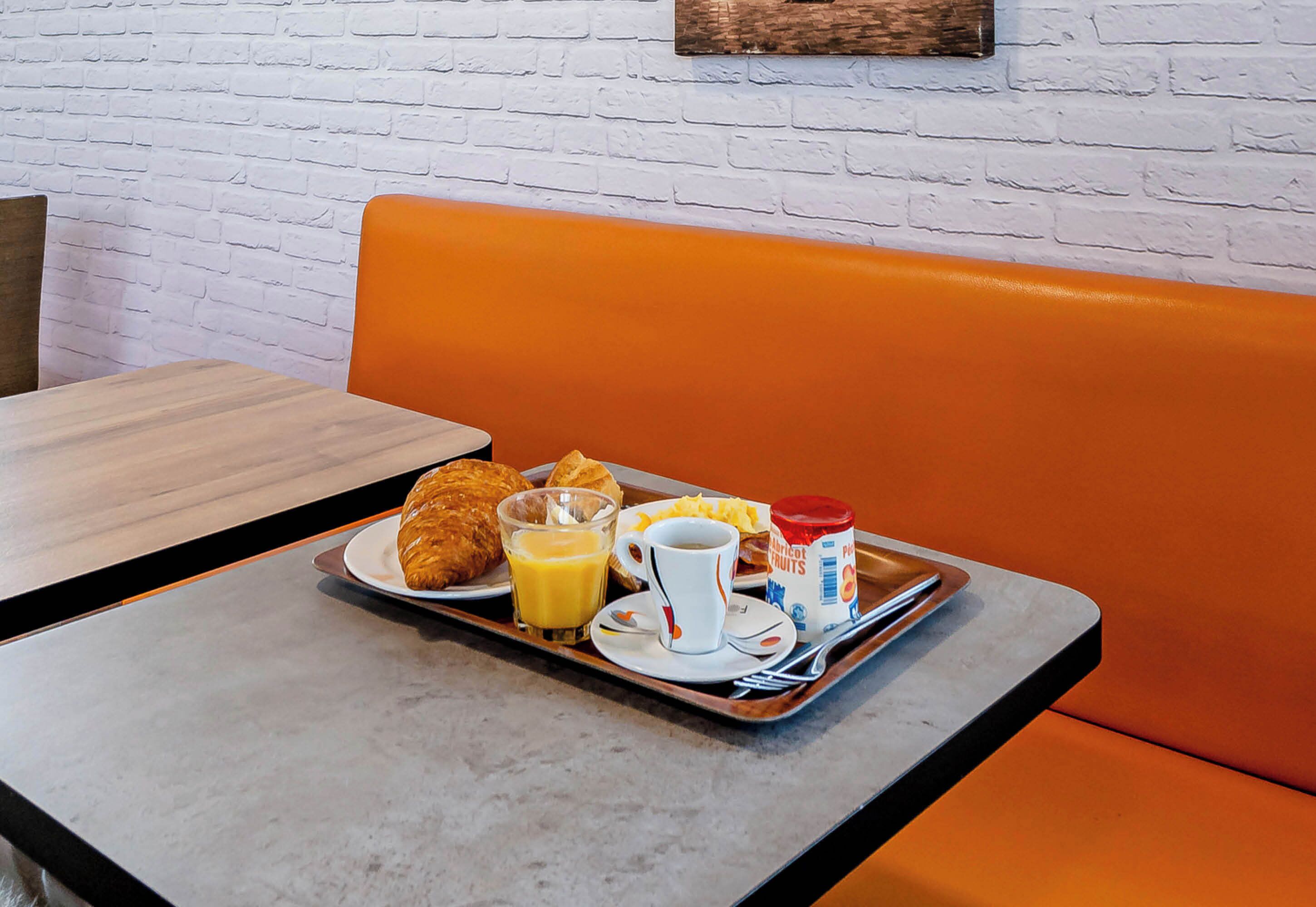 Bandeja de desayuno con croissants, jugo de naranja, una taza y un envase de yogur con fruta sobre una mesa con un banco acolchado naranja.