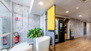 Eine moderne Lobby mit Holzboden, weißen Stühlen, einer Topfpflanze, einem Feuerlöscher und einem Getränkeautomaten.