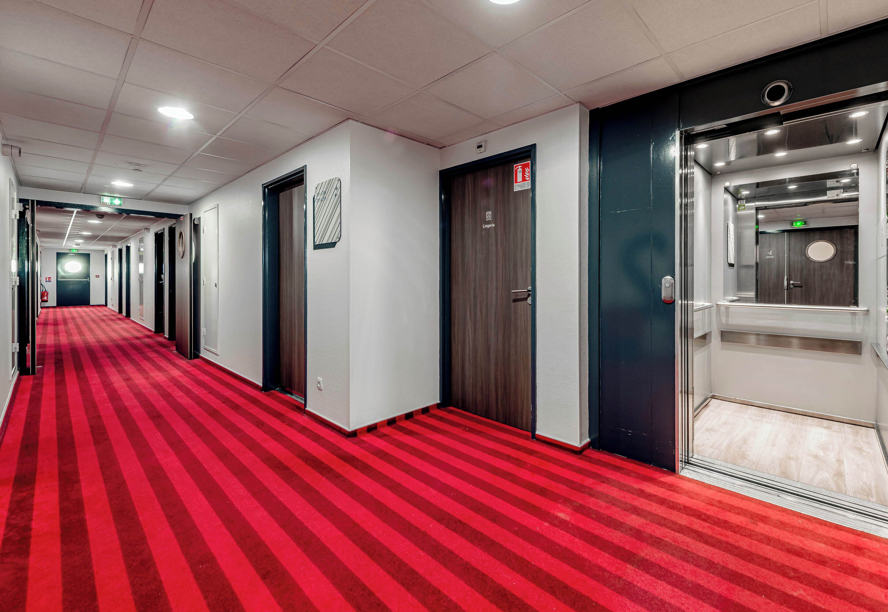 Een hotelgang met rode tapijttegels, deuren en een lift.
