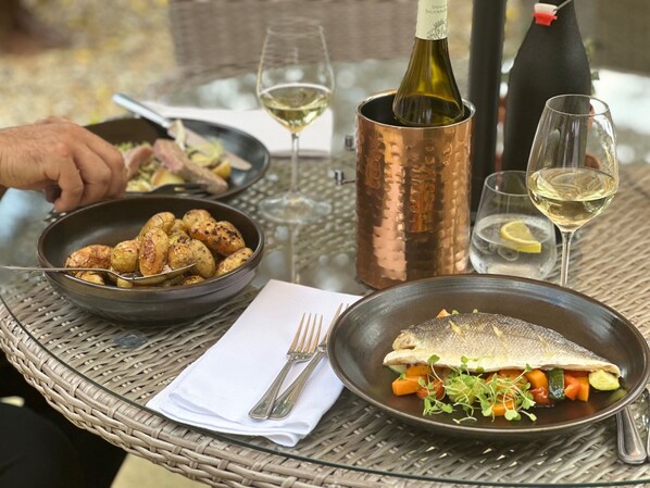 Una mesa de comedor con un plato de papas asadas, pescado y verduras, acompañado de copas y botellas de vino.