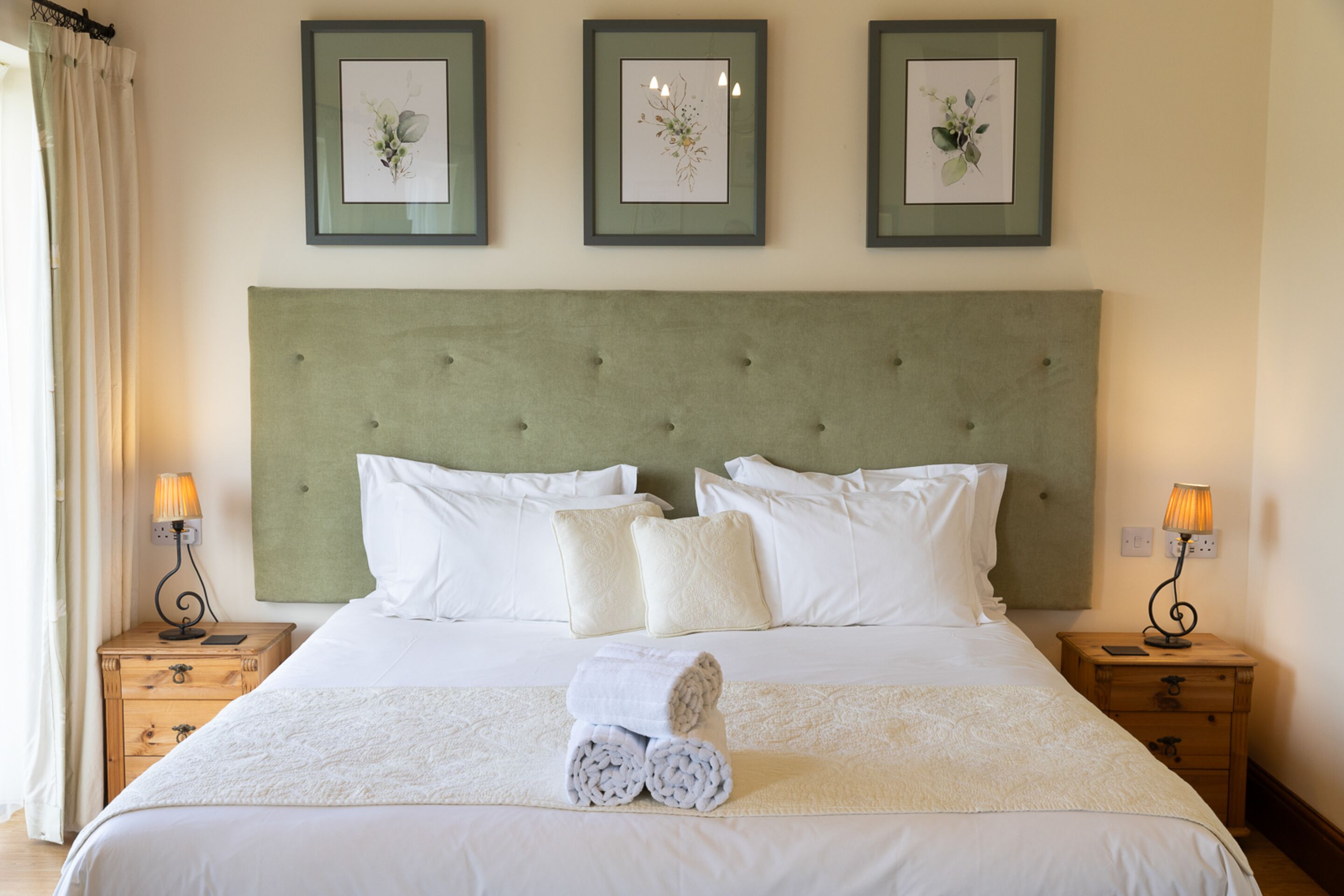 Un lit bien fait, avec du linge de lit blanc, deux tables de chevet avec des lampes, et trois œuvres d’art florales encadrées accrochées au mur.