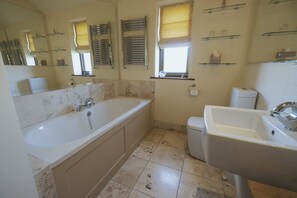 A bathroom with a bathtub, a sink, and a toilet. There are shelves with toiletries and a window with blinds.