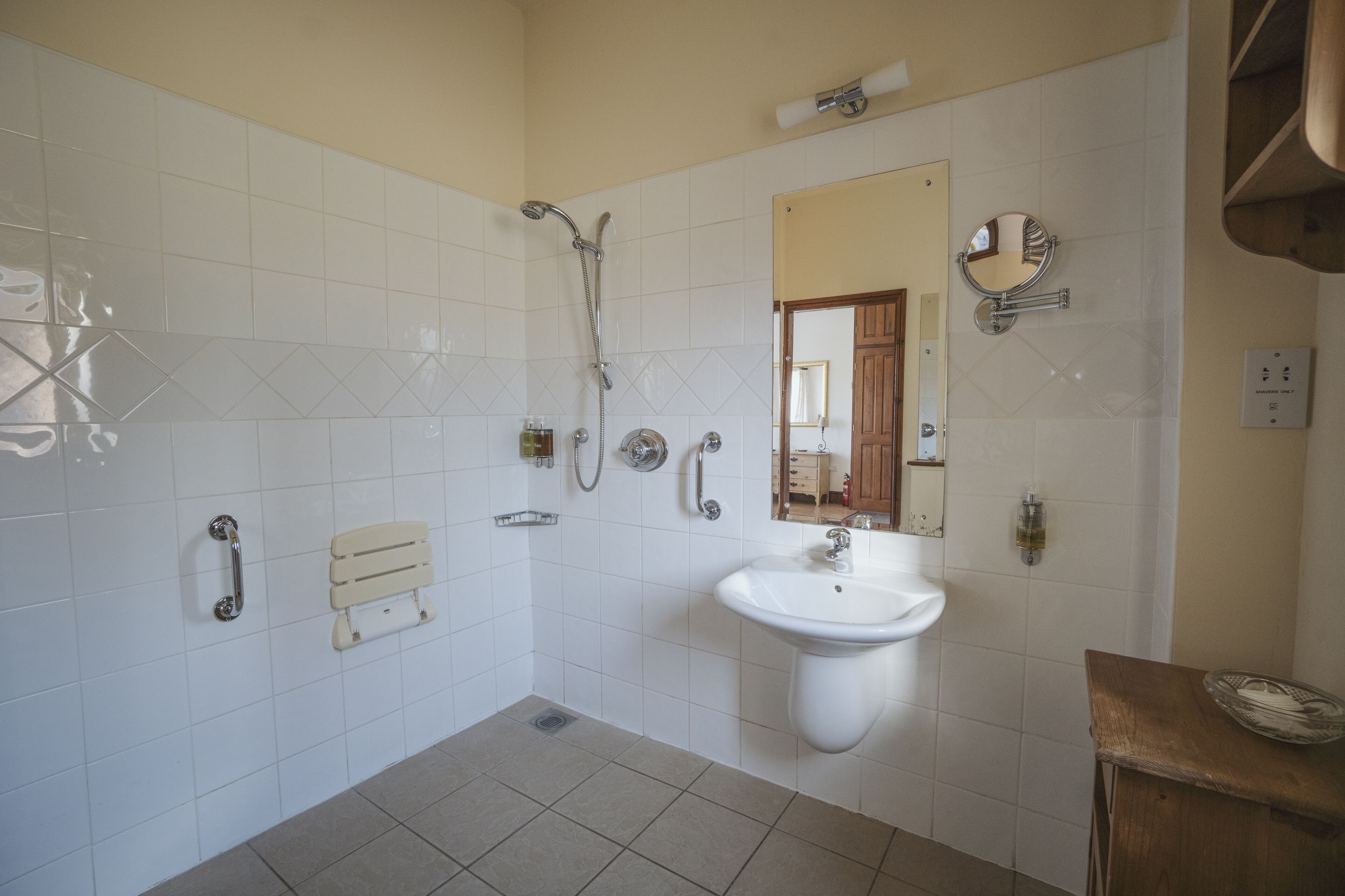 Casa de banho com azulejos brancos, uma pia, um espelho, um chuveiro e um banco de madeira.