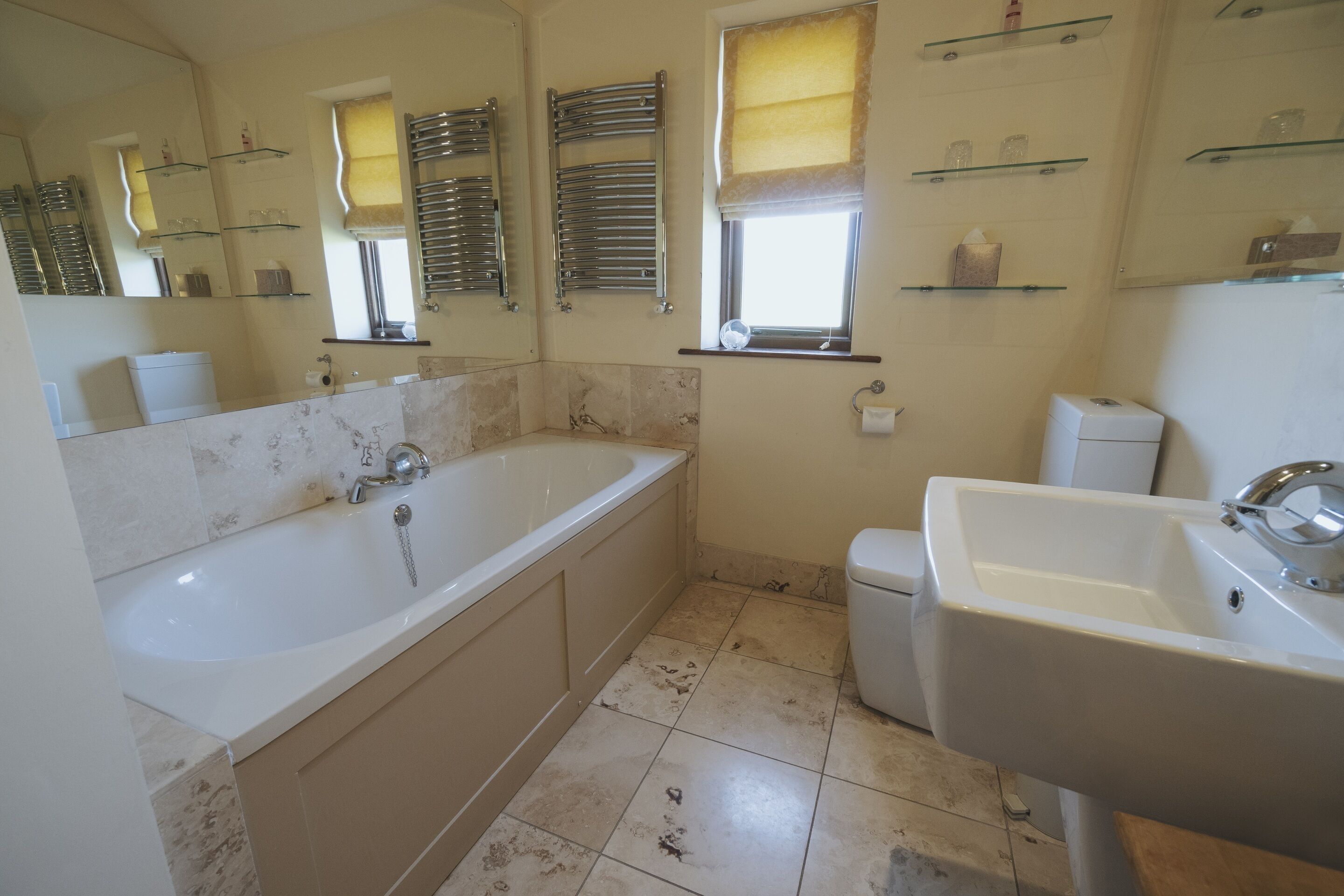 A bathroom with a bathtub, a sink, and a toilet. There are shelves with toiletries and a window with a towel rack.
