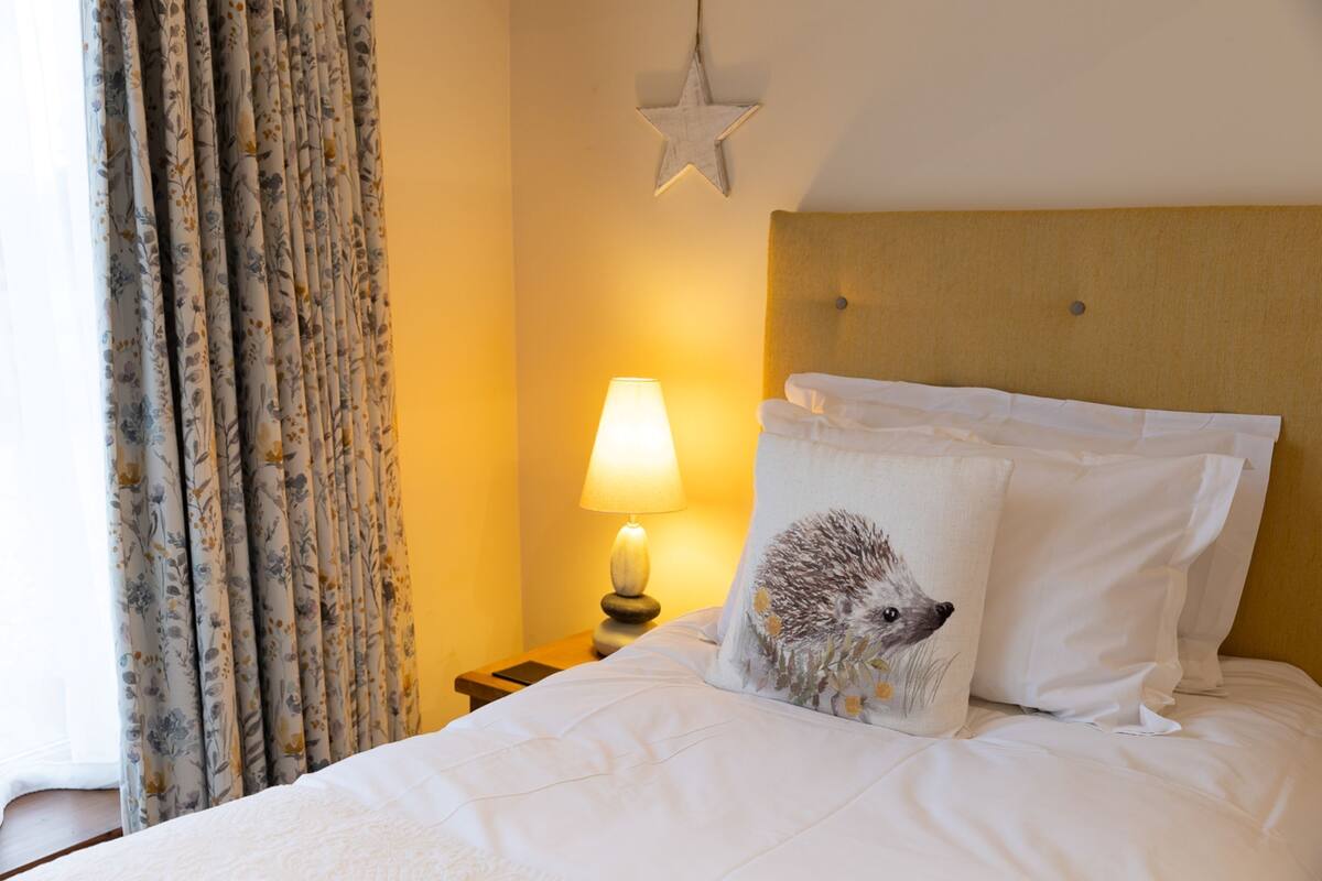 Uma cama bem arrumada com roupa de cama branca, um travesseiro fofo com desenho de ouriço, um abajur e um enfeite de parede em formato de estrela.