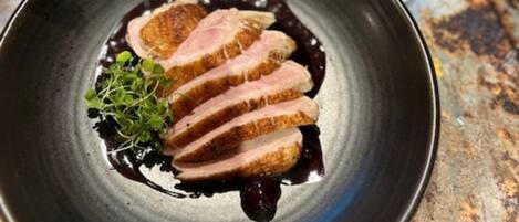 Une assiette de viande tranchée nappée d’une sauce foncée, accompagnée de légumes verts, un verre de vin rouge et, au fond, un récipient en métal avec un couvercle bleu.
