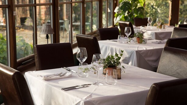A dining area with tables set for two, featuring white tablecloths, wine glasses, and potted plants.