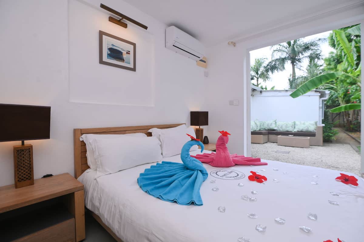 A hotel room with a bed, bedside tables, a wall-mounted air conditioner, a framed picture, and a view of a garden with palm trees.