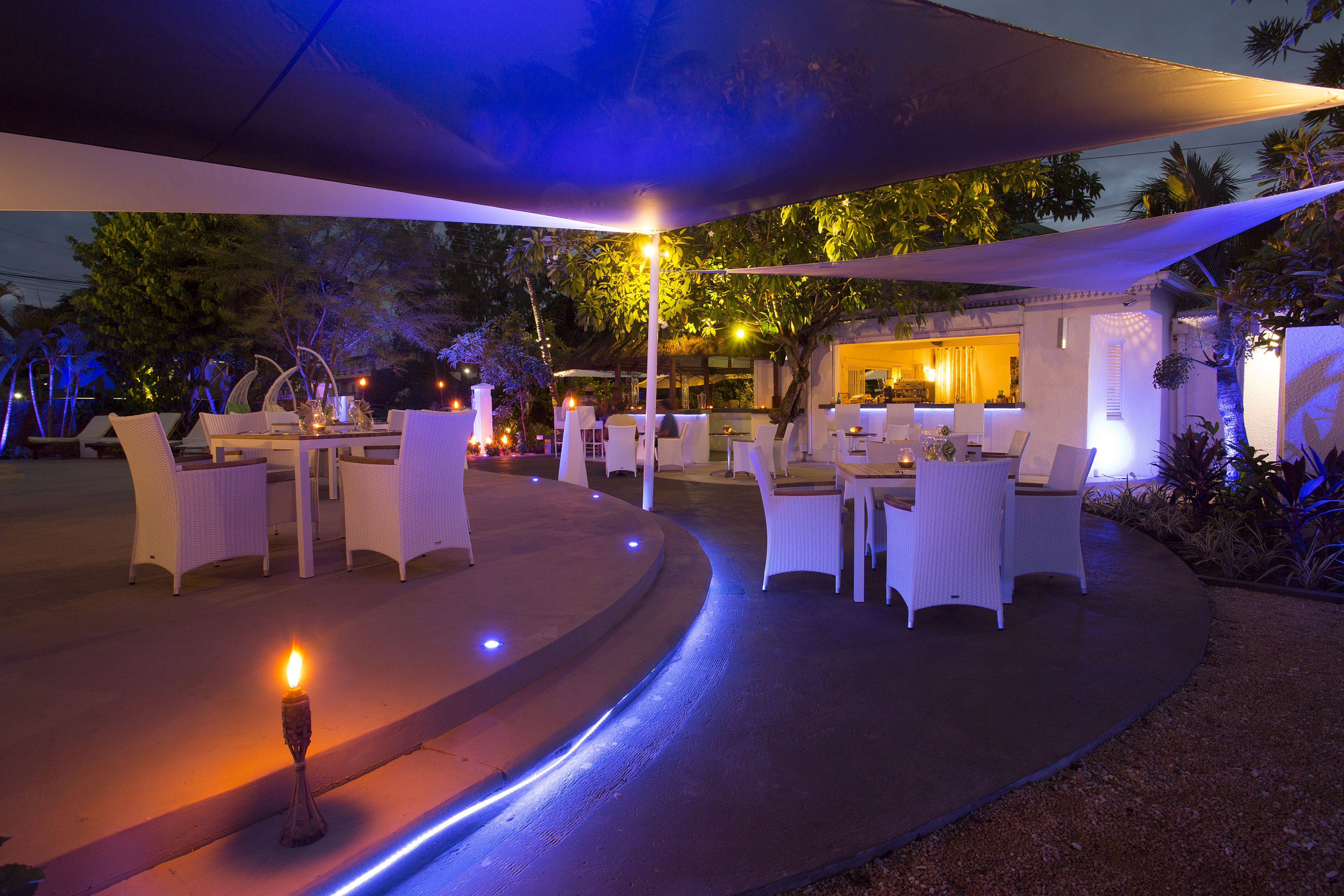 An outdoor dining area with white wicker chairs and tables, under a shaded canopy, with a lit candle on the ground.