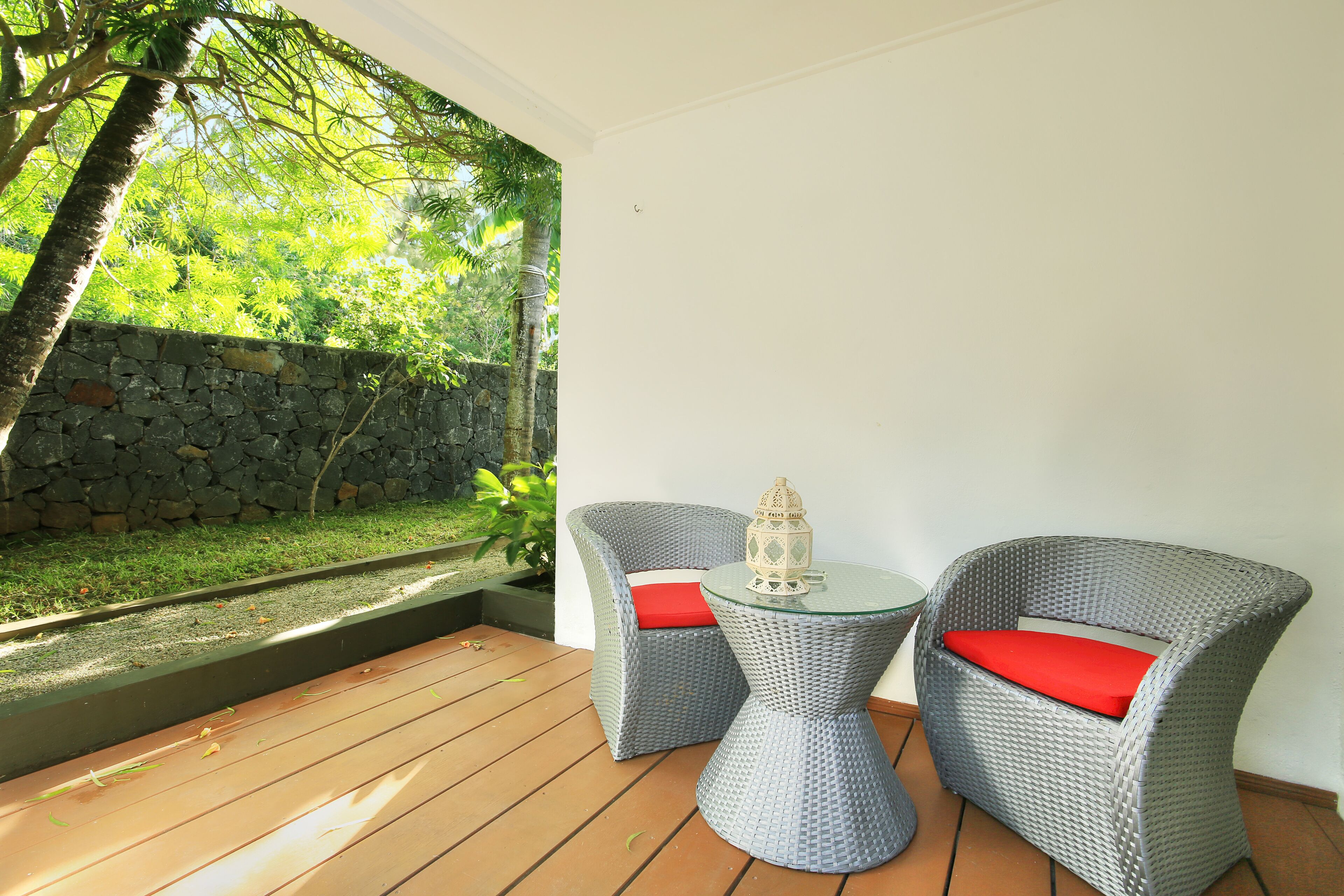 Een terras met rieten meubels en een glazen tafel, met uitzicht op een tuin met een stenen muur en bomen.