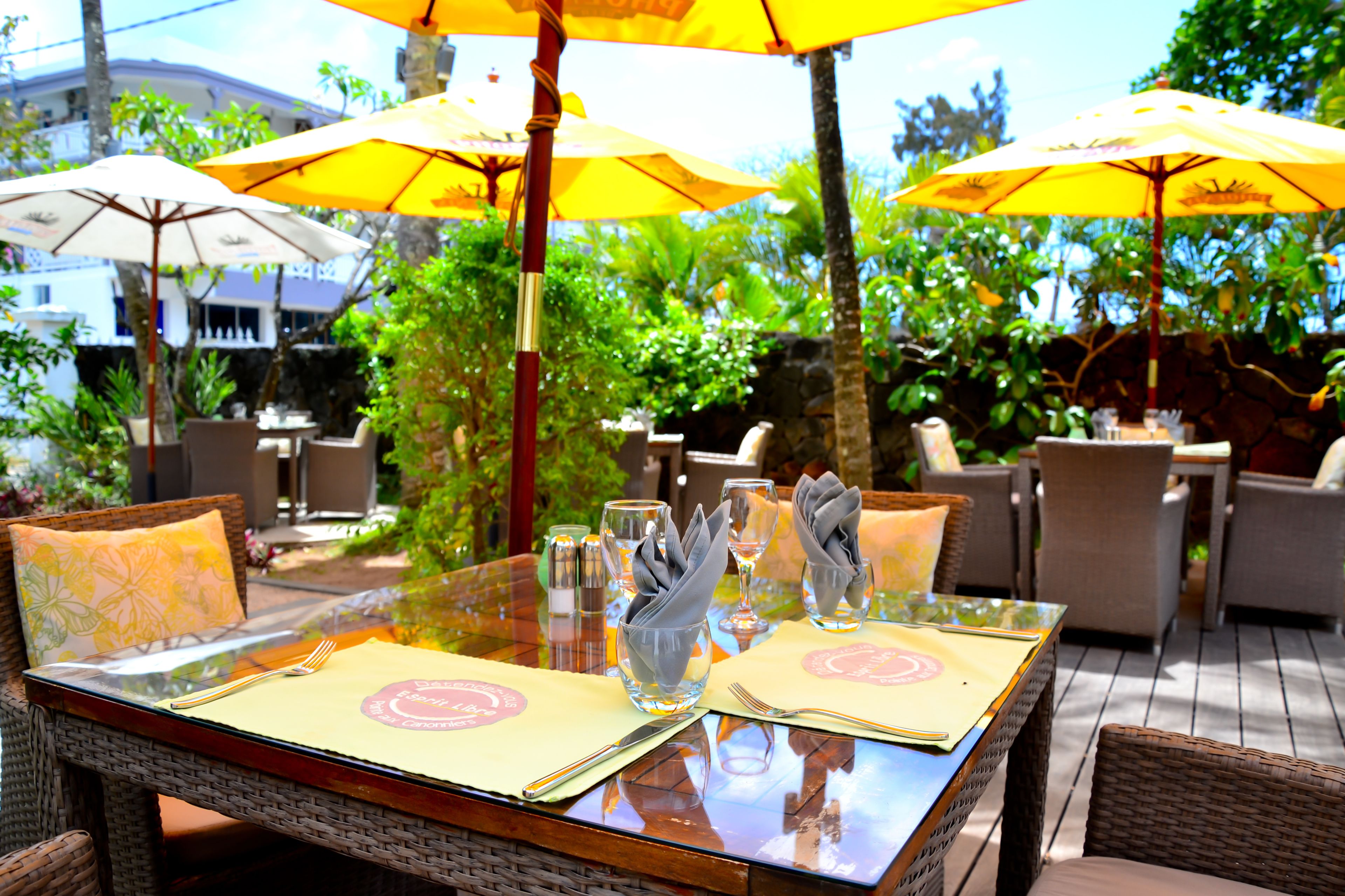 An outdoor dining area with wicker furniture, yellow umbrellas, and glass tables.
