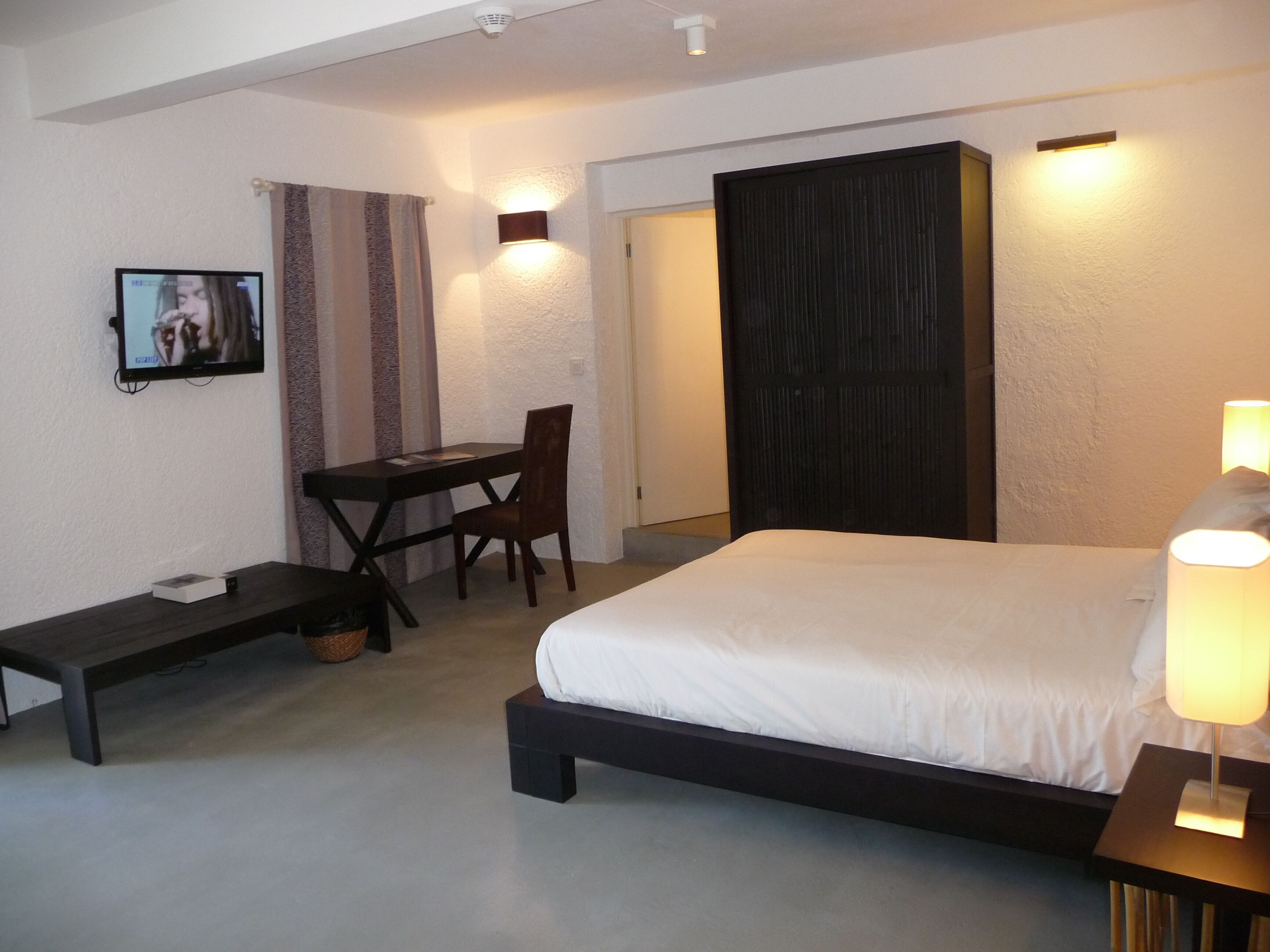 Een slaapkamer met een bed, nachtkastje, lamp, bureau, stoel en een televisie aan de muur.