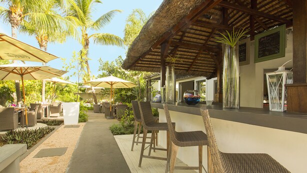 An outdoor bar area with wicker chairs and umbrellas, surrounded by palm trees and greenery.