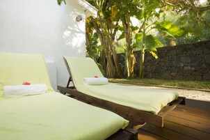 Dos sillones de madera con cojines verdes y toallas blancas, ubicados al aire libre frente a un muro de piedra y abundante vegetación.