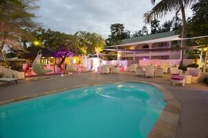 Piscina con sillas de descanso y sombrillas, rodeada por un edificio con techo verde y un patio cubierto.
