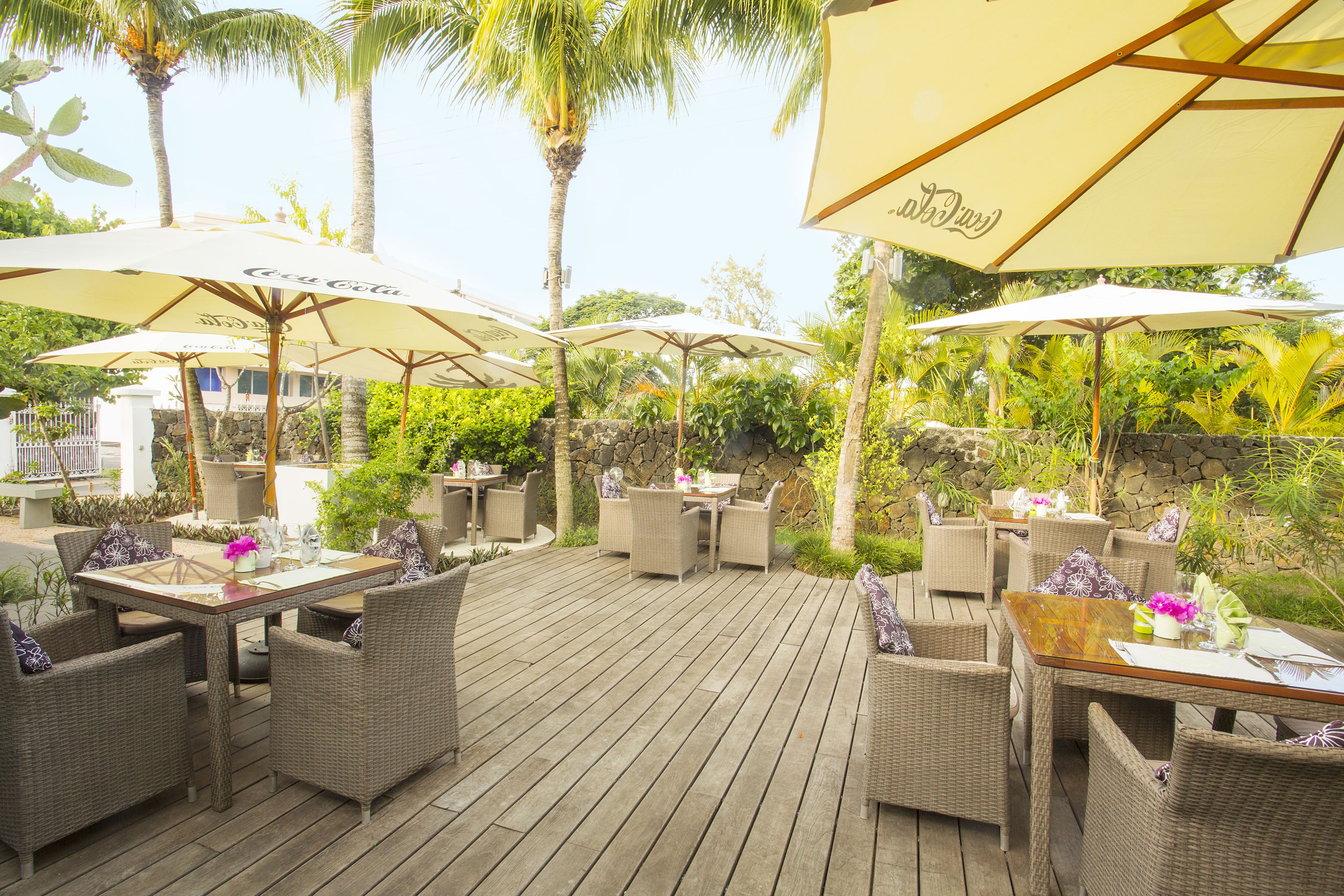 Een buiten eethoek met rieten meubels en grote parasols, omgeven door tropische planten en palmbomen.