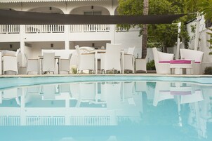 Un área junto a la piscina con muebles de mimbre blanco y una piscina cristalina.