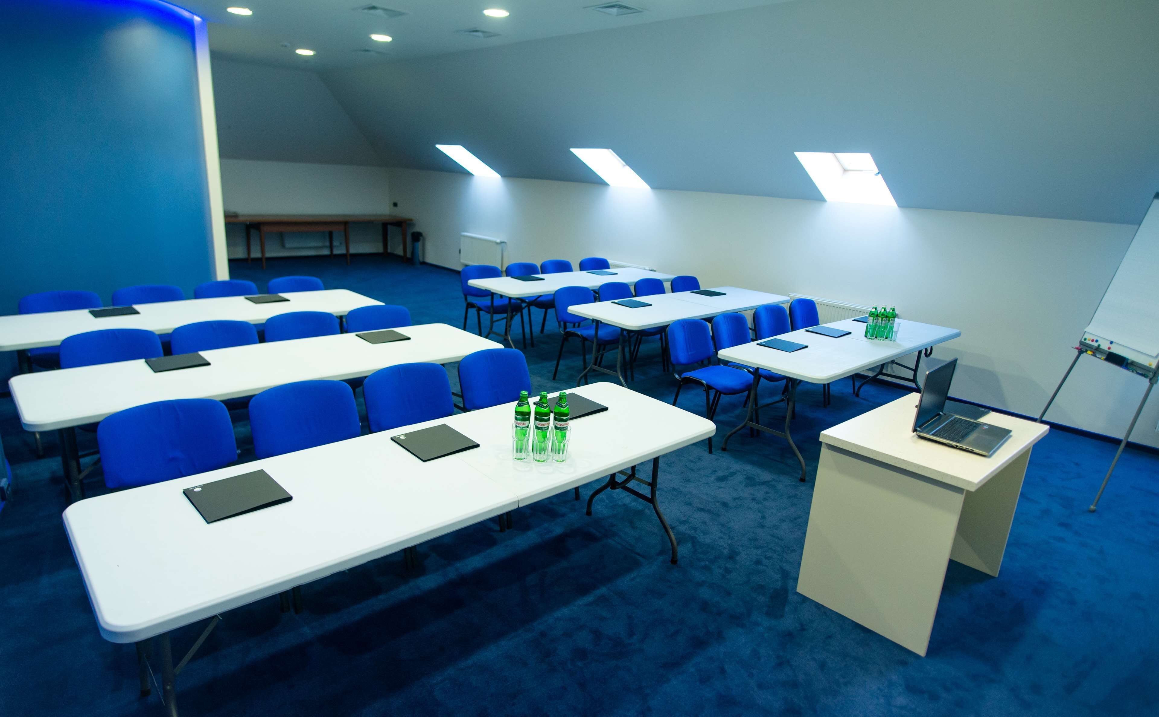 A conference room with rows of blue chairs, white tables, and a podium with a laptop.