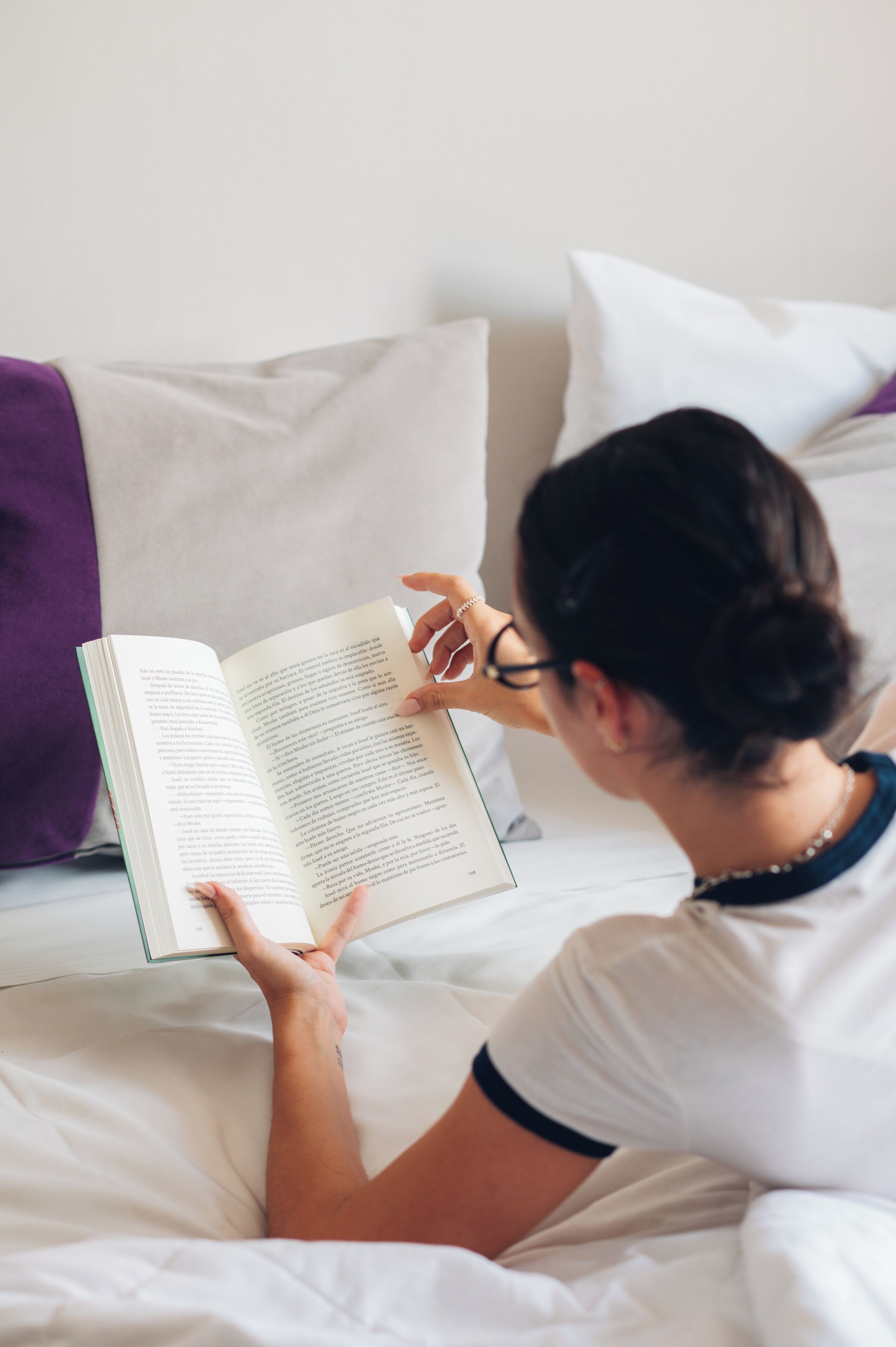 A person reading a book on a bed with white bedding and a purple pillow.
