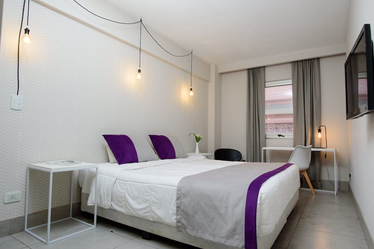 A hotel room with a bed, a desk, and a chair. There is a window with curtains, a wall-mounted TV, and a wall socket.
