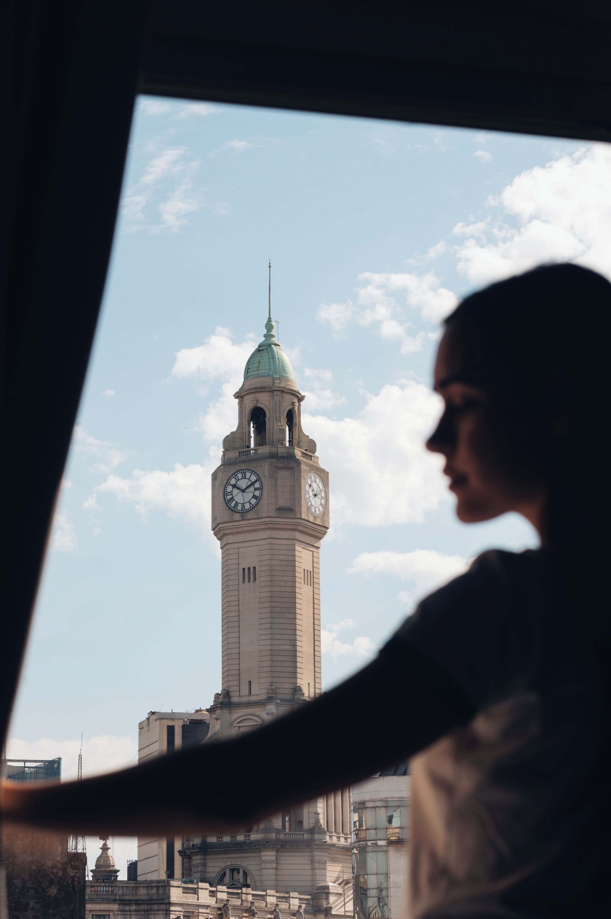 A view of a clock tower with a dome and a spire, seen through a window with a partial view of a person.