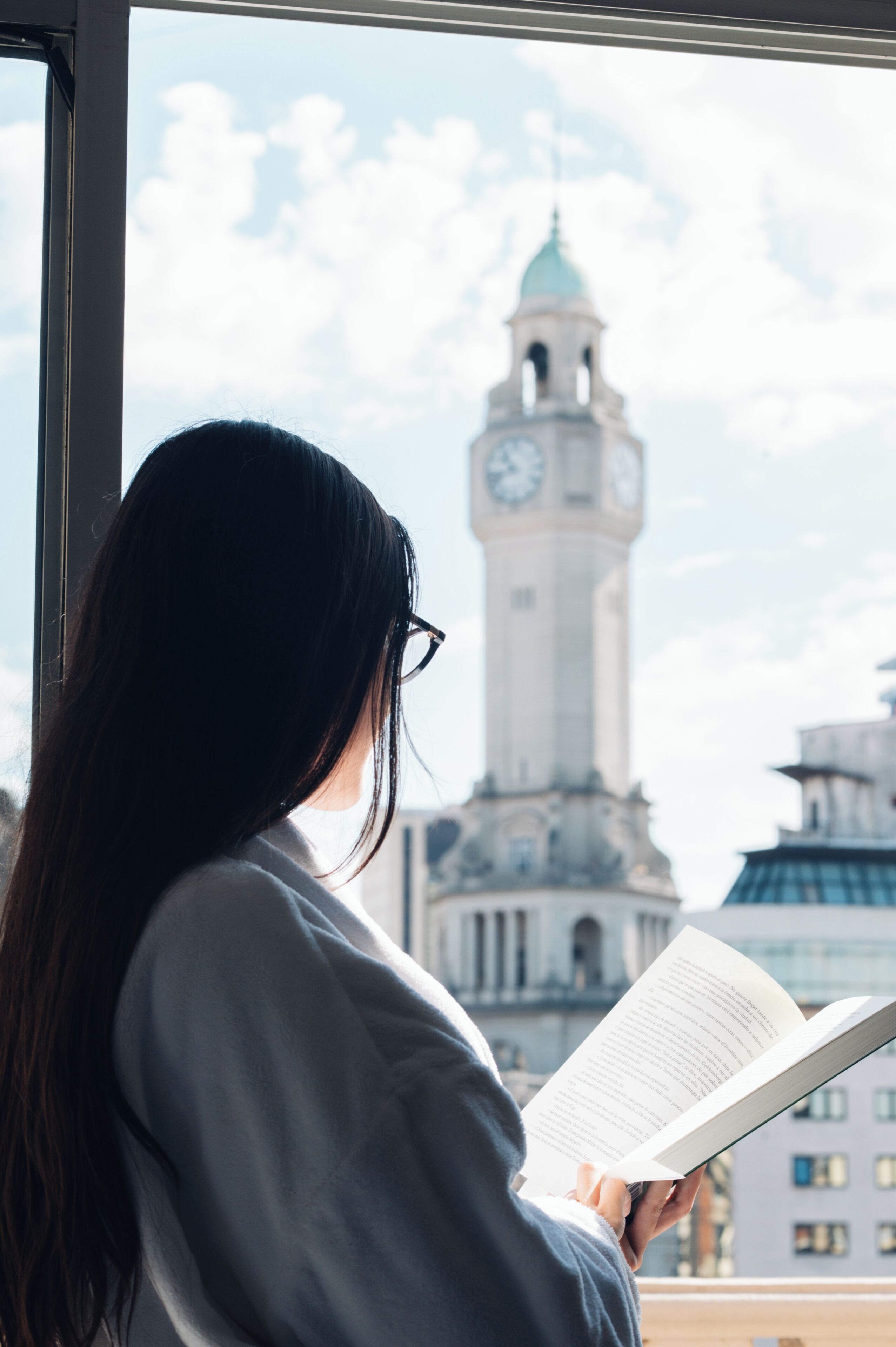 Una persona llegint un llibre a la finestra amb vista a una torre del rellotge.