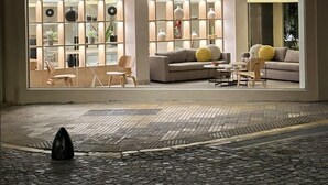 A modern hotel entrance with a glass facade, interior seating, and a cobblestone ground.