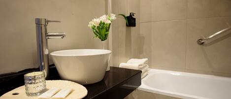 Un baño con un lavamanos grande de color blanco, una encimera negra, un candelabro de cristal, un florero con flores blancas, un dispensador de jabón negro y una bañera blanca.