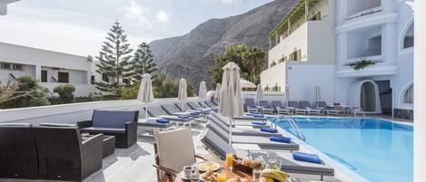 A rooftop pool area with lounge chairs and umbrellas, a dining table with breakfast, and a mountain backdrop.