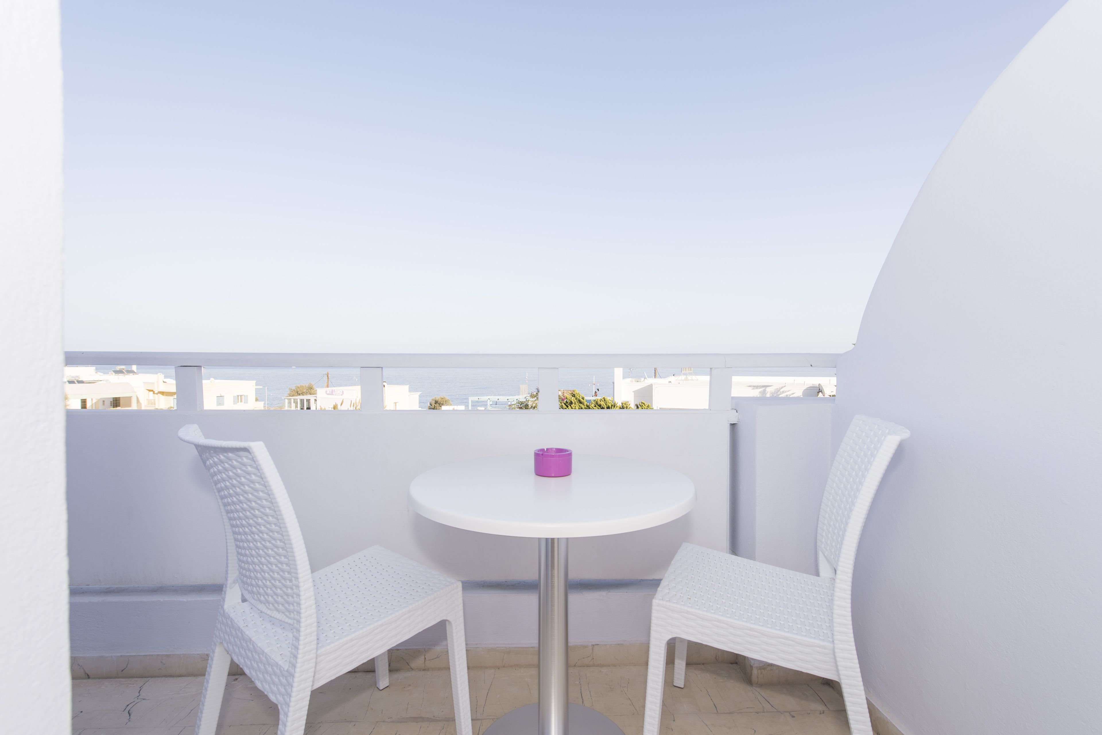 A small balcony with a white table and two white chairs, a purple object on the table, and a view of buildings in the background.