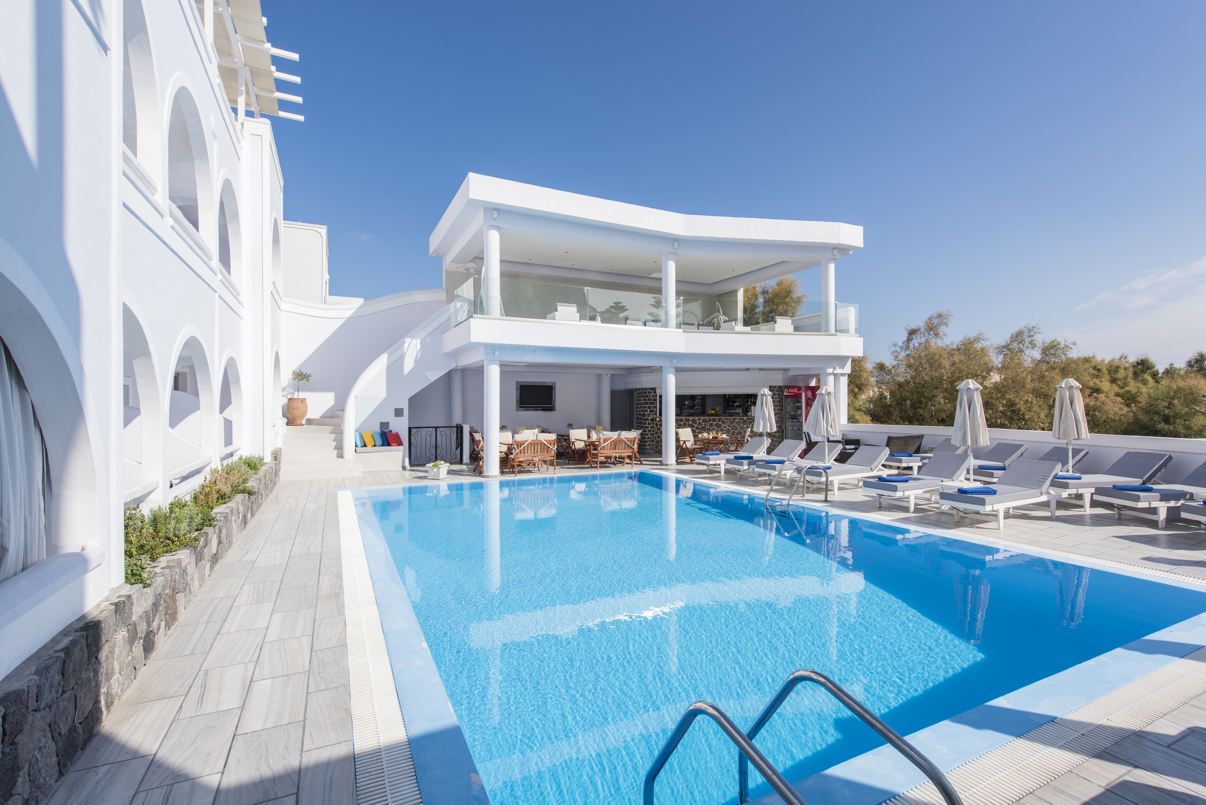 Un área de piscina moderna con piscina azul cristalina, sillas de descanso y un edificio de dos pisos con balcones.