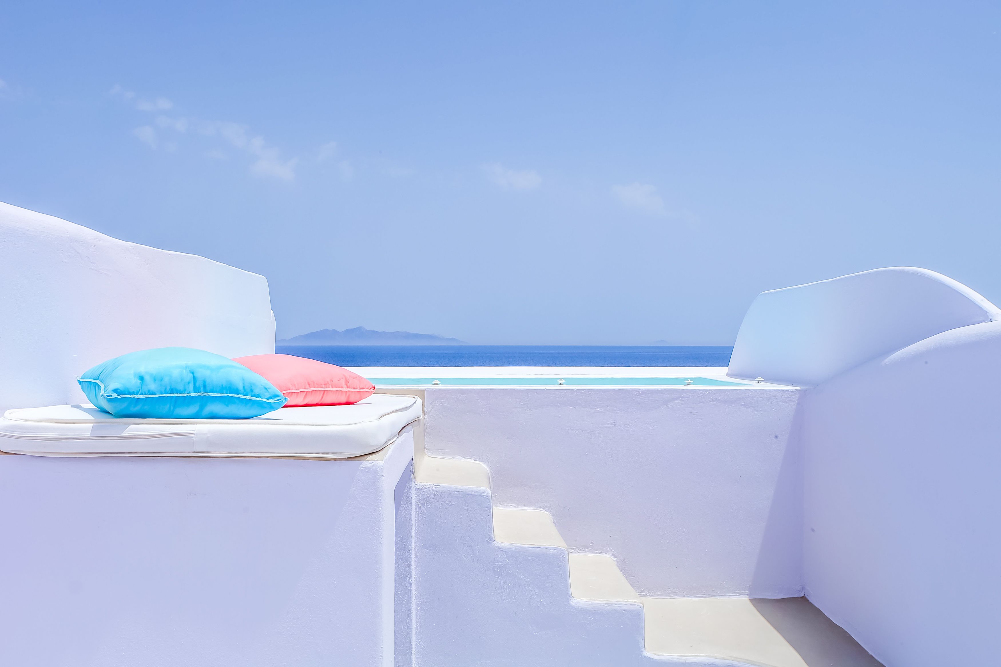 Un área de estar exterior blanca con escalones que bajan a una piscina, con vistas al mar azul cristalino y montañas lejanas.
