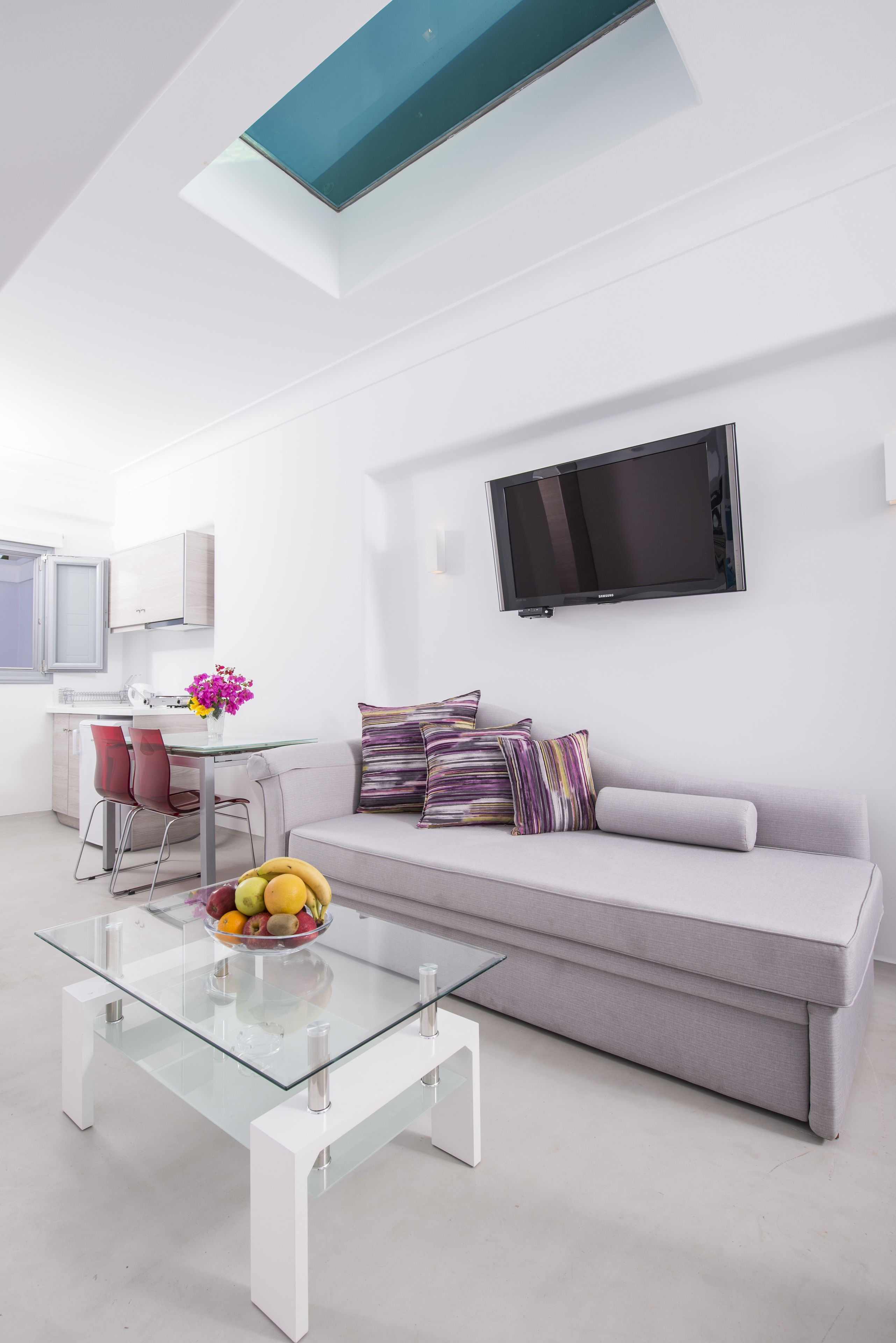 A modern living room with a glass coffee table, a grey sofa, and a flat-screen TV mounted on the wall.